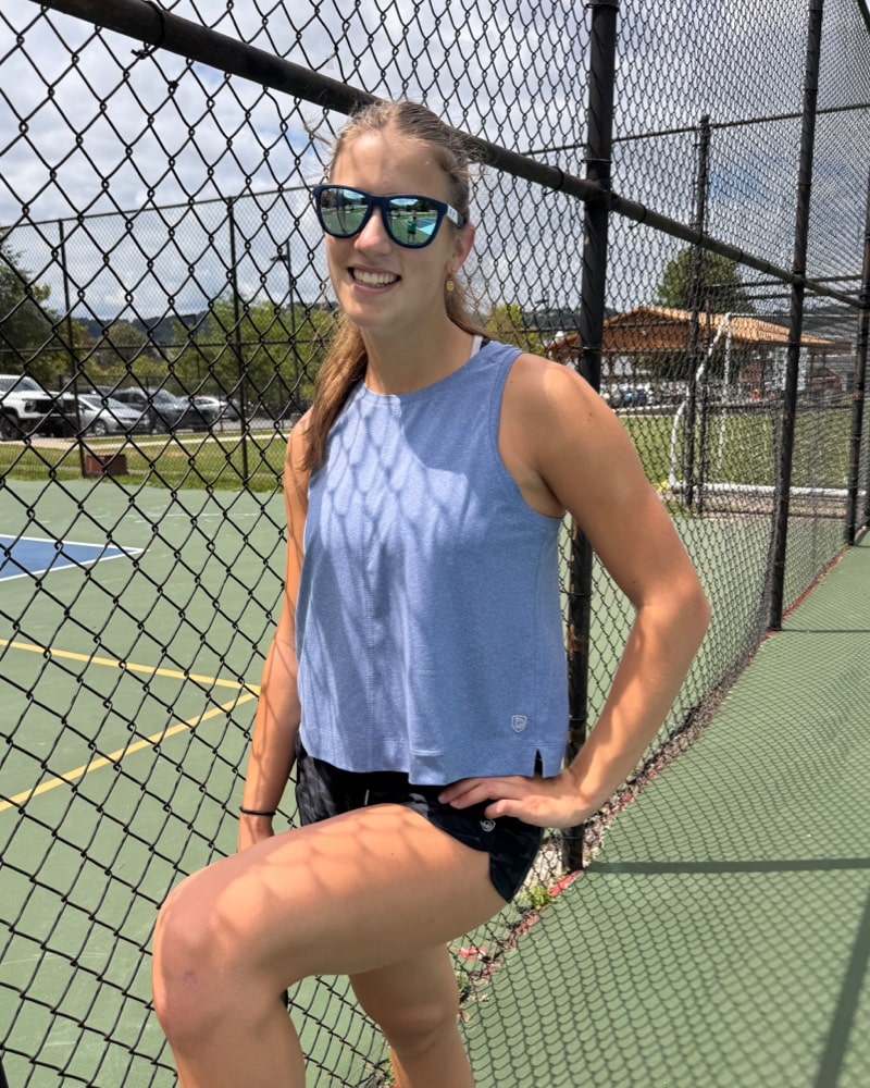 Blue Boxy Crop Tank, front three-quarter view beside a fence at an outdoor court