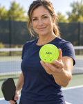 AI-assisted pickleball scene of one adult woman holding a single lime-yellow ARTI Outdoor Pickleball before play. Original supplier photos show product details.