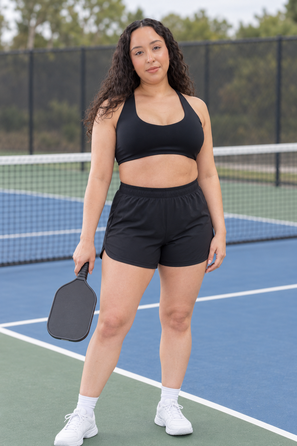 AI-assisted pickleball scene: one adult woman wearing BRUSHED commit sports bra in Black — post point pause.