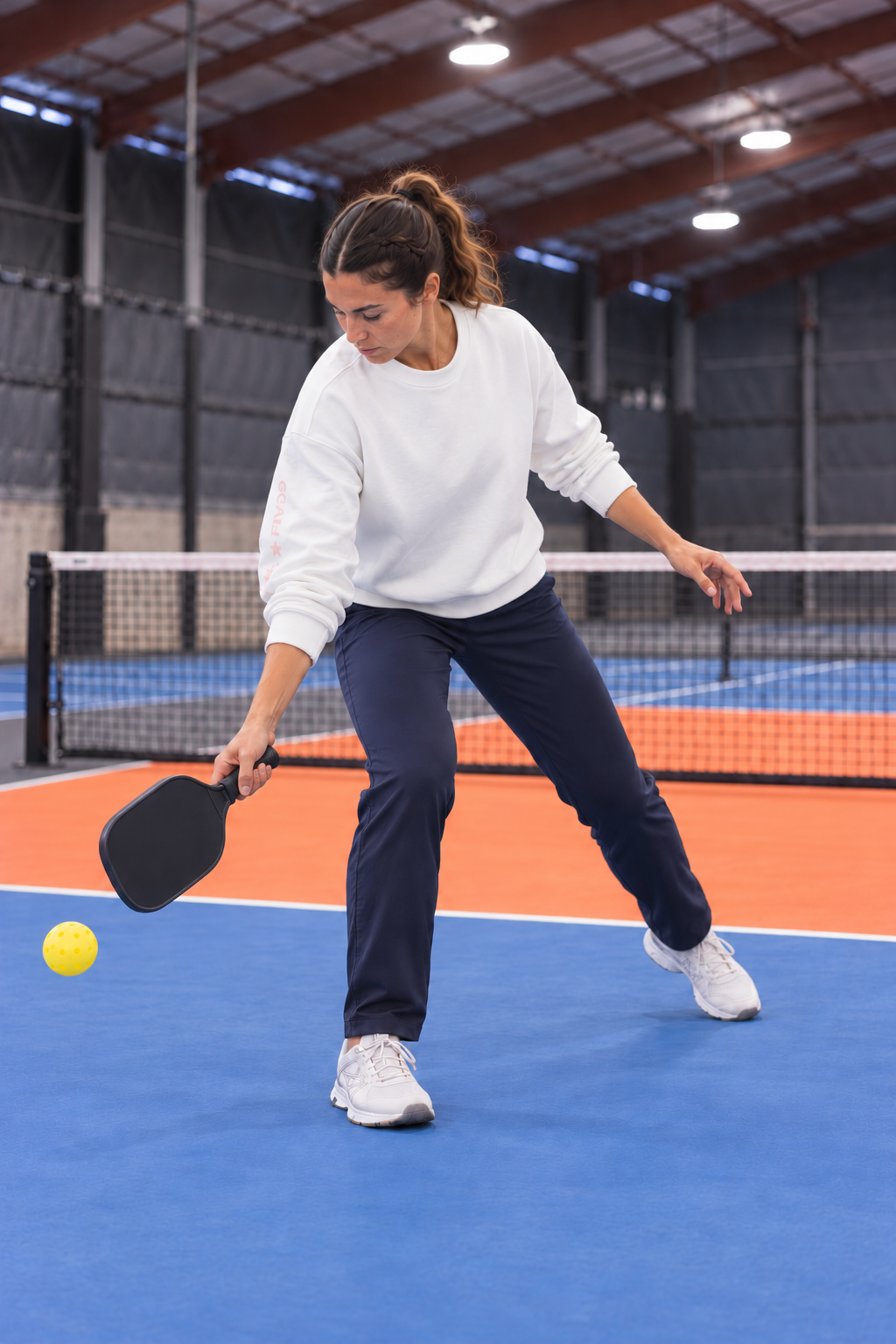 AI-assisted: same adult woman in the White Freasy Crew preparing a low pickleball return on a Portland-region-inspired indoor court.