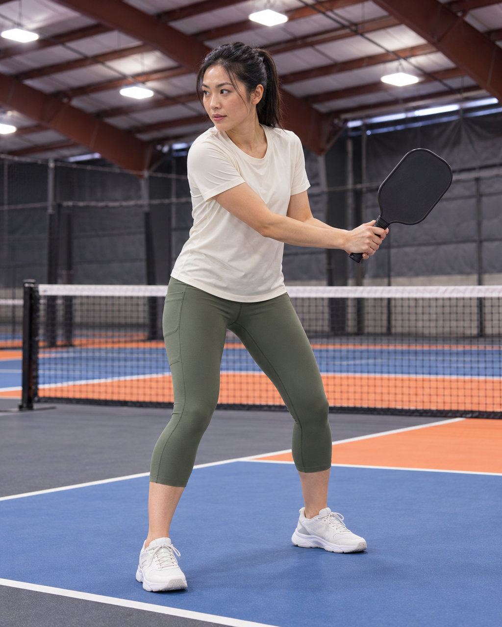 AI-assisted editorial image of an adult woman preparing a compact backhand on a Portland-region-inspired indoor pickleball court wearing Go Again cropped leggings in Dark Moss. Venue-inspired generated scene.