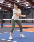 AI-assisted editorial image of an adult woman preparing a compact backhand on a Portland-region-inspired indoor pickleball court wearing Go Again cropped leggings in Dark Moss. Venue-inspired generated scene.