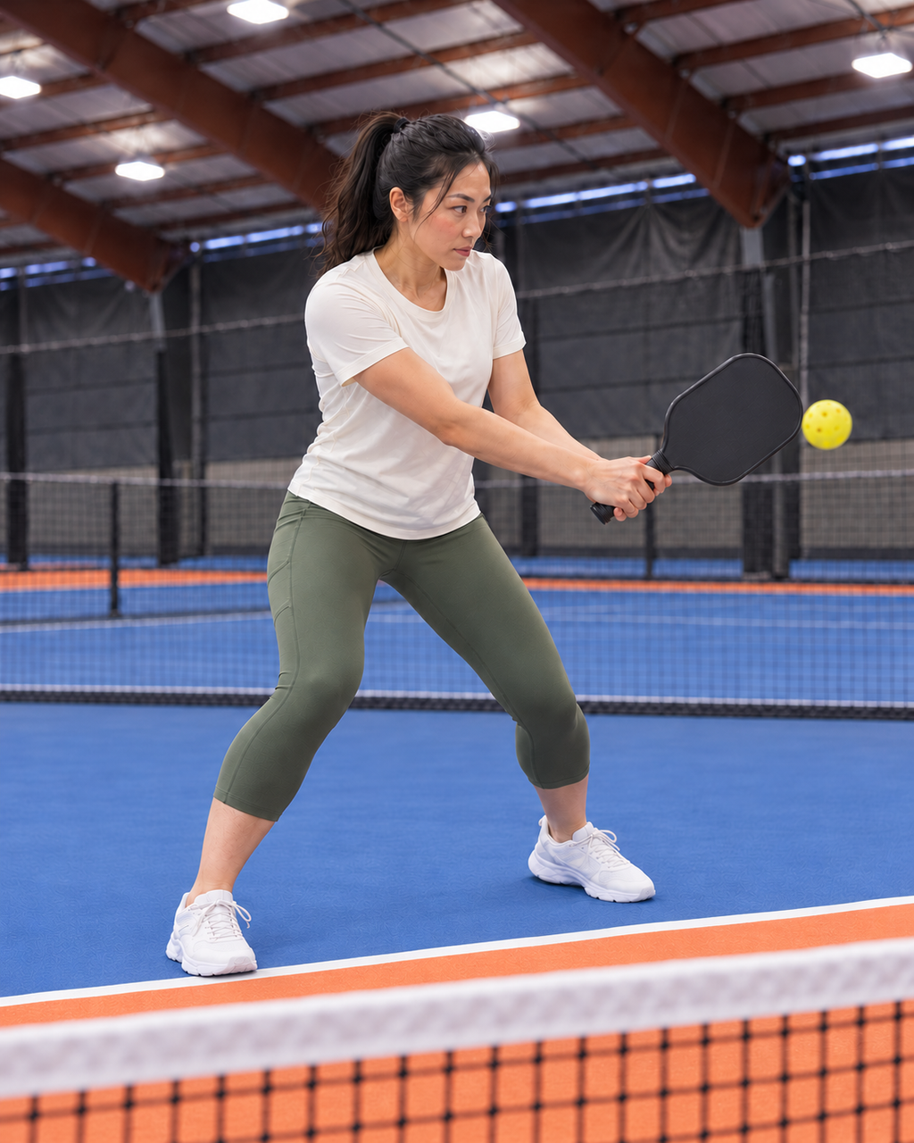 AI-assisted editorial image of an adult woman in a compact two-handed backhand volley wearing Go Again cropped leggings in Dark Moss on a Portland-region-inspired indoor pickleball court. Venue-inspired generated scene.