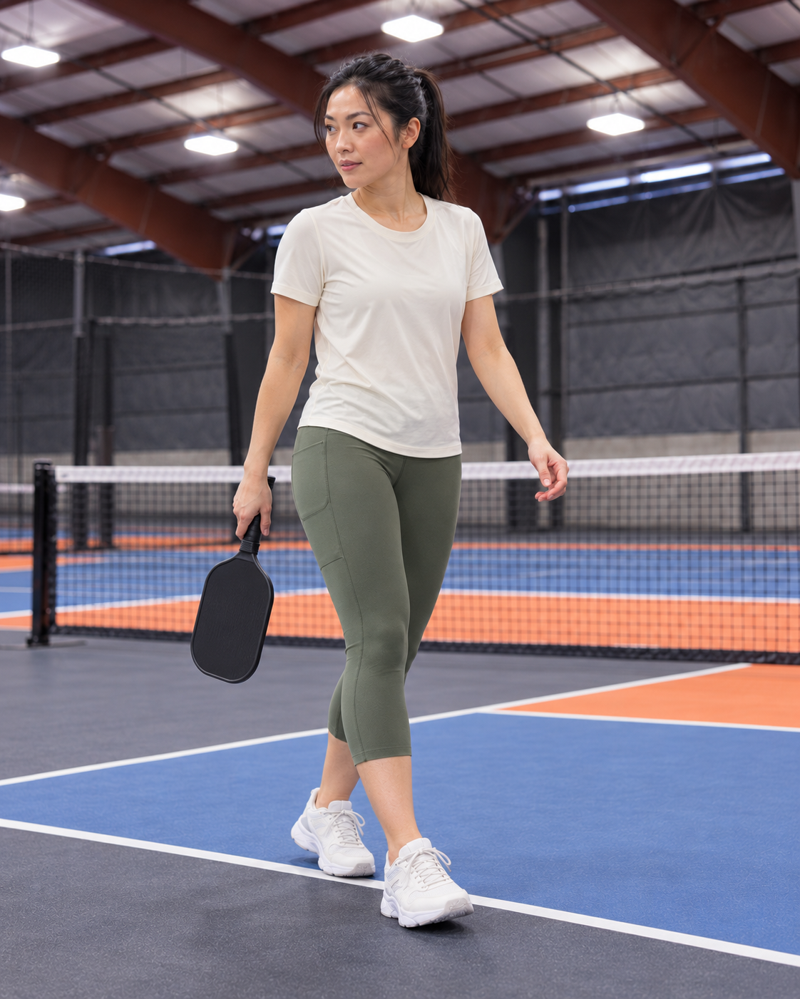 AI-assisted editorial image of an adult woman in walking between points wearing Go Again cropped leggings in Dark Moss on a Portland-region-inspired indoor pickleball court. Venue-inspired generated scene.